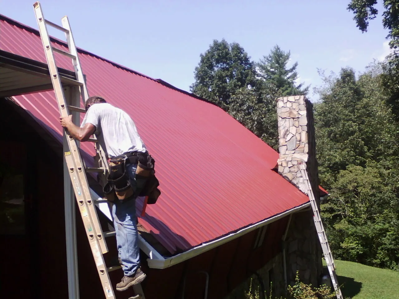 Roofer on a ladder beside a red metal roof and stone chimney — Soffit & Fascia Repair by Roofing Brothers in Wilkeson